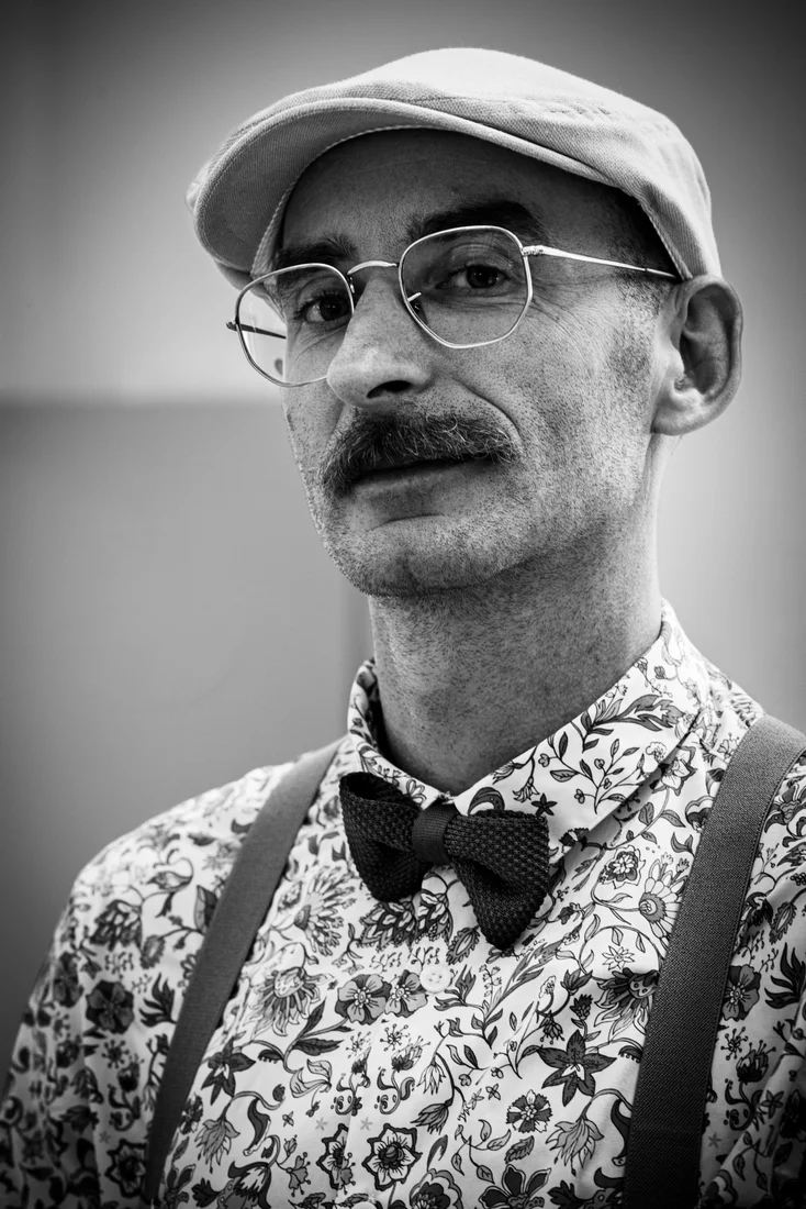 Portrait en noir et blanc d’un homme portant une casquette, des lunettes et un nœud papillon.
